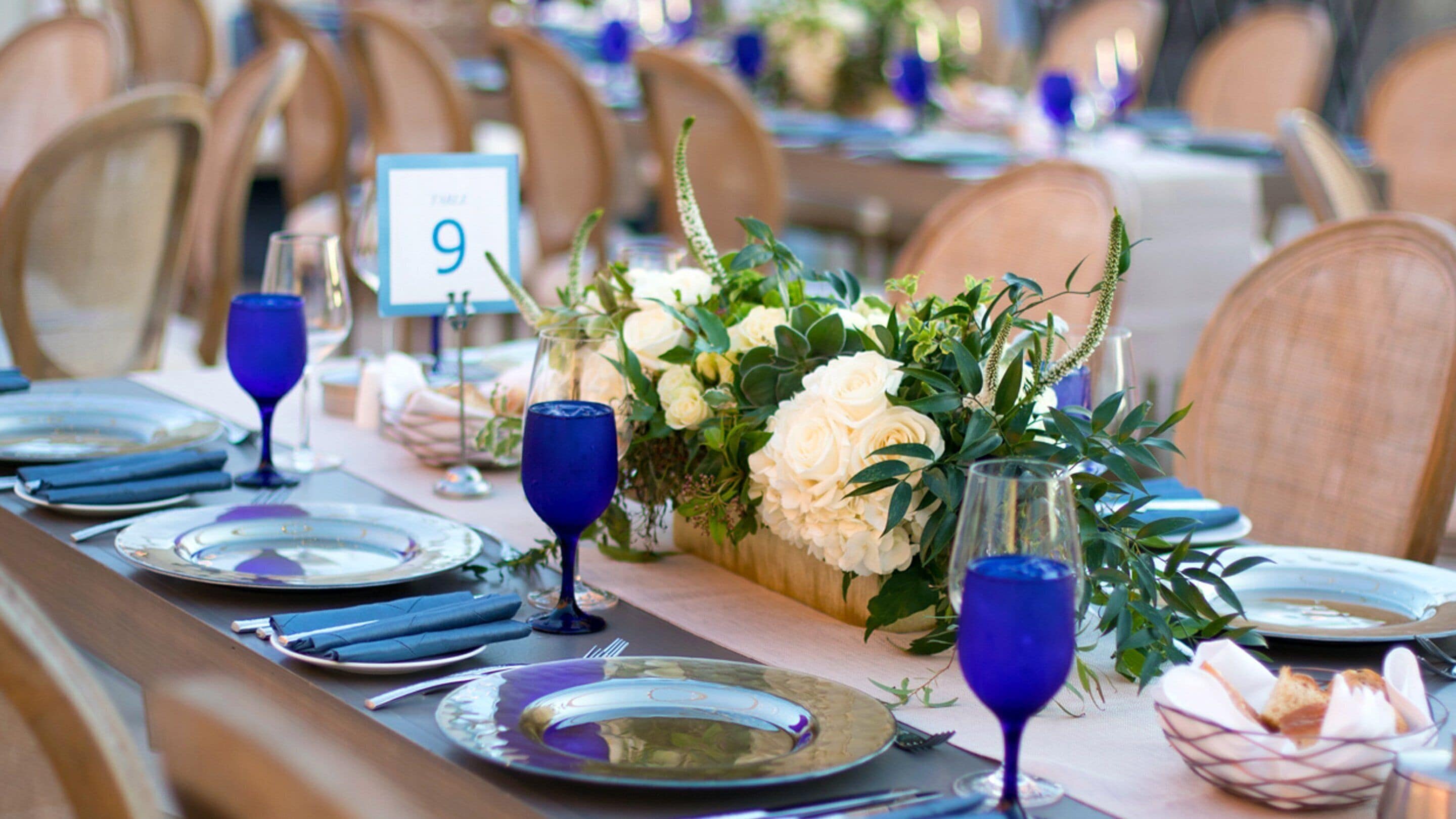 Mesa dispuesta para un evento formal, con manteles y sillas numeradas, copas de vidrio azul y centros de mesa florales.