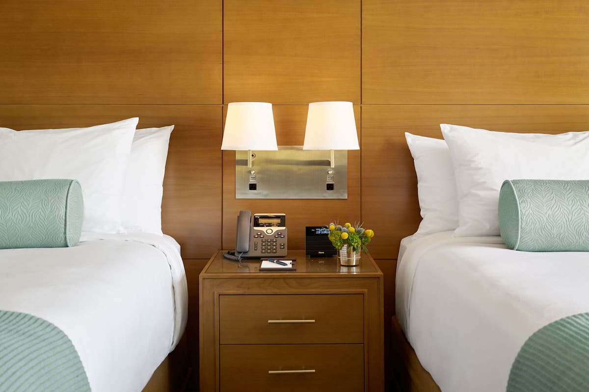 A hotel room with two beds, a bedside table with a lamp, a telephone, and a small vase with flowers.