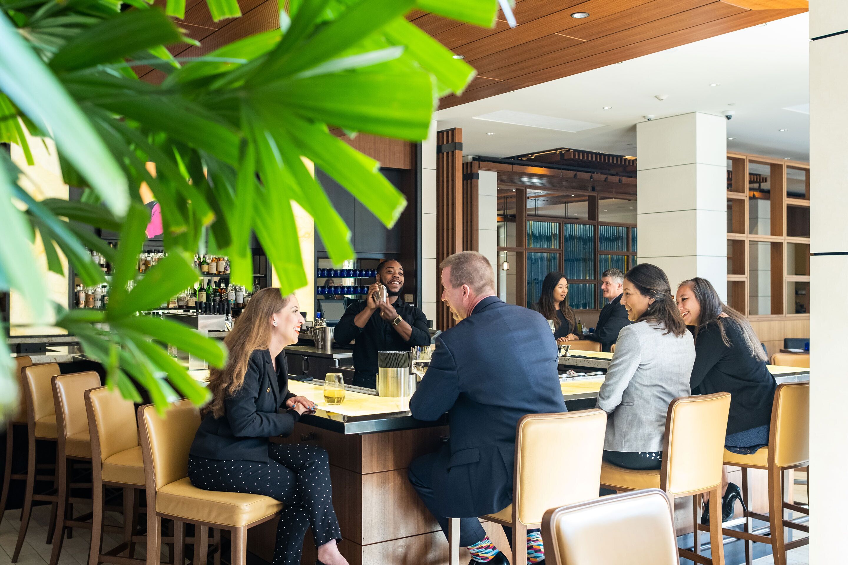 A bar area with several people seated at tables, a bartender behind the bar, and a modern interior design with wooden elements and plants.