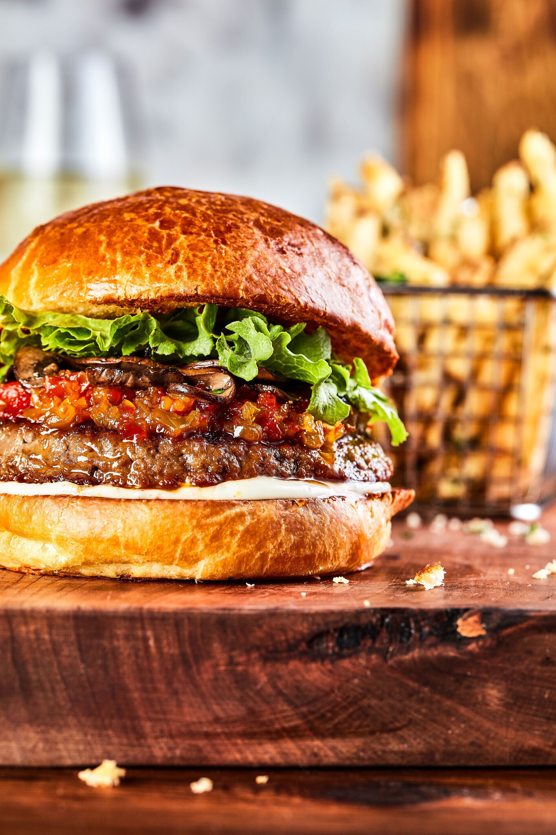 A burger with lettuce and sauce on a bun, placed on a wooden surface with a basket of fries in the background.