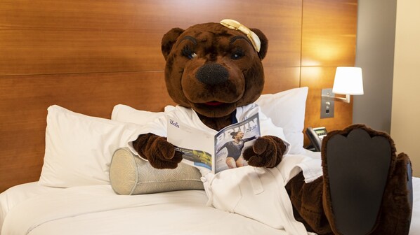 A plush bear mascot lying on a bed with a magazine, a pillow, and a nightstand with a lamp.