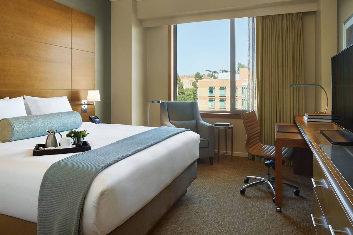 A hotel room with a large bed, a desk with a flat-screen TV, a chair, a small table, and a window with a view of buildings.