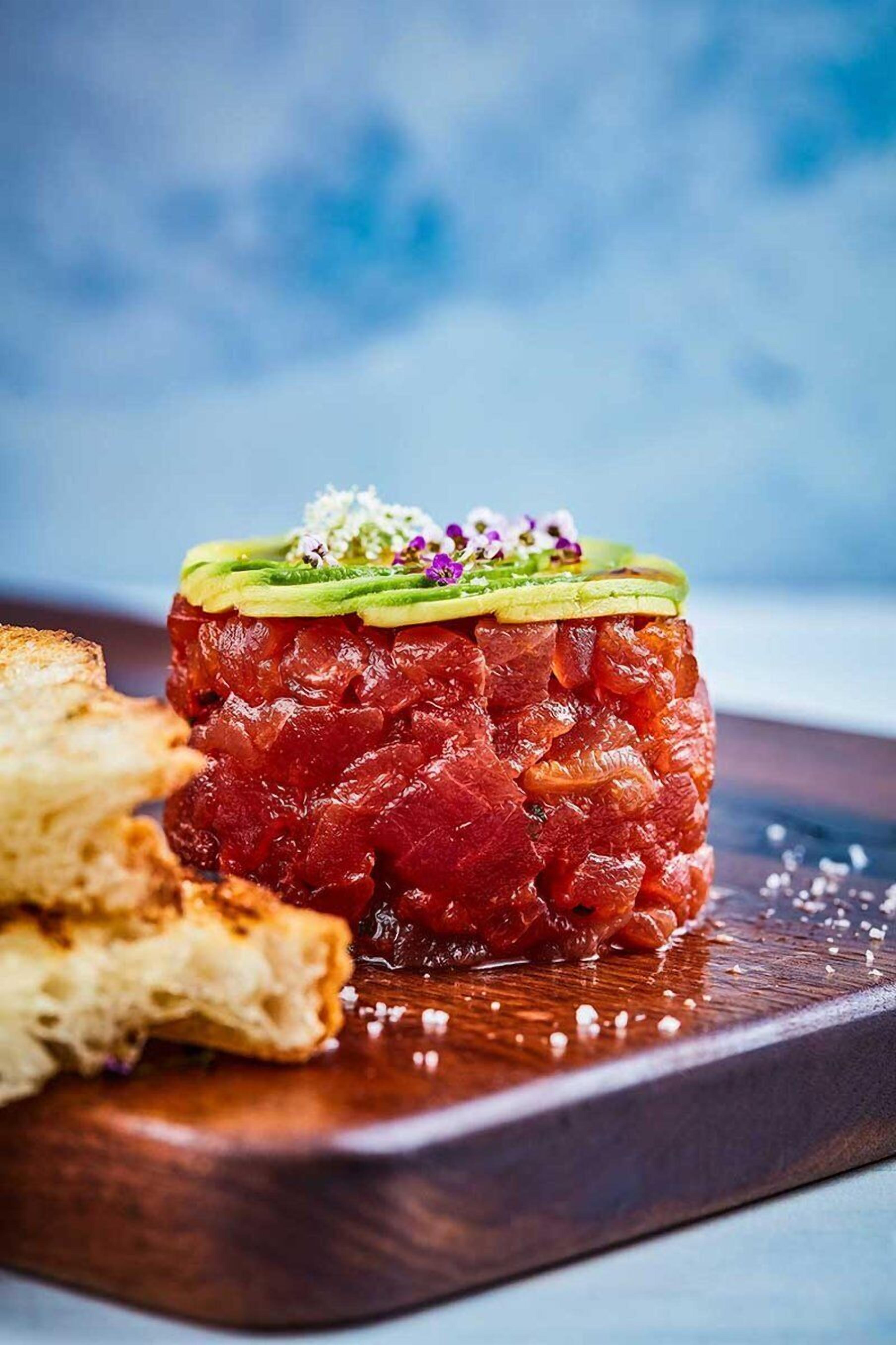 Un plat de tartare de thon cru, garni de tranches d’avocat et de fleurs comestibles, présenté sur une planche en bois.