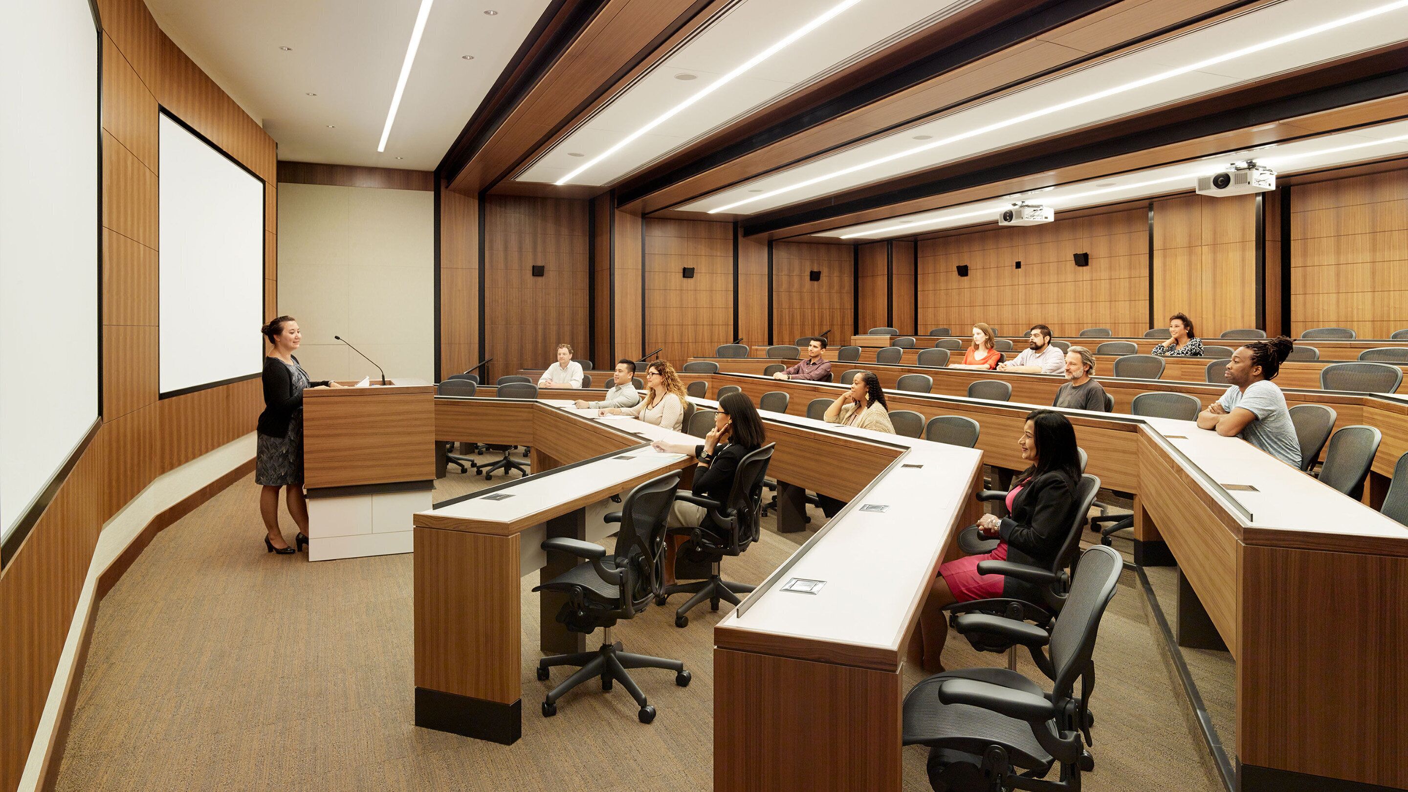 A conference room with a speaker at the podium, attendees seated at desks, and a projector screen.