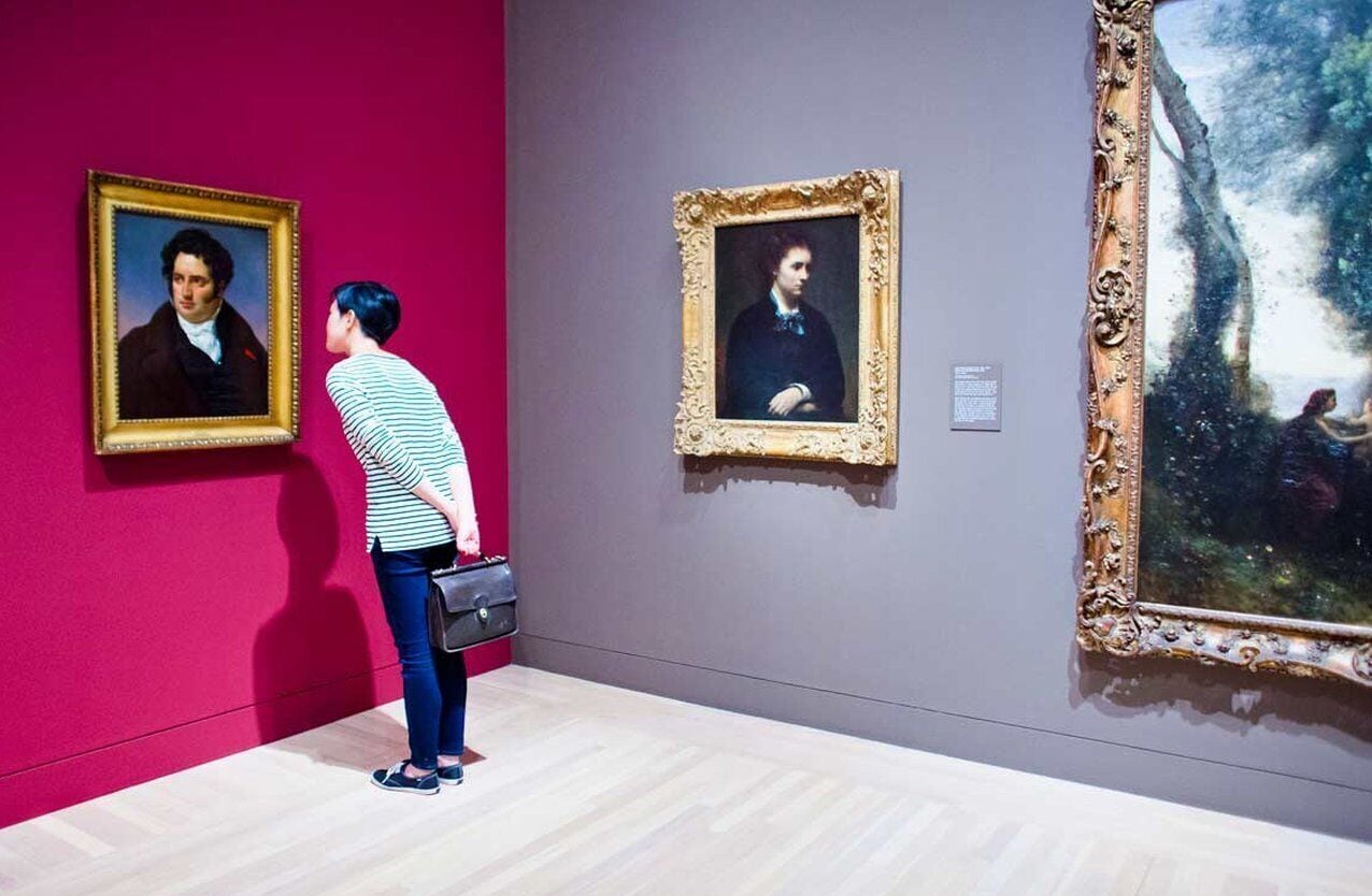 Una persona observa pinturas en una galería de arte con una pared rosa y un cuadro grande enmarcado a la derecha.