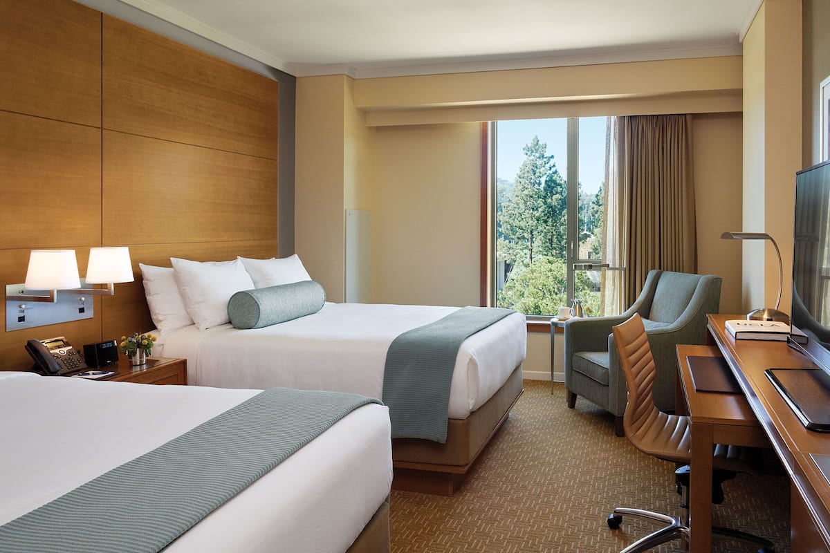 A hotel room with two beds, a desk, a chair, a TV, and a window with a view of trees.