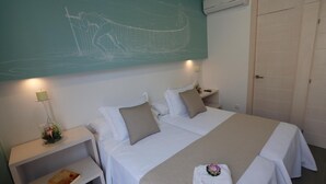 Habitación de hotel con dos camas, un mural en la pared, una mesita de noche con una lámpara y una mesita junto a la cama con un libro.