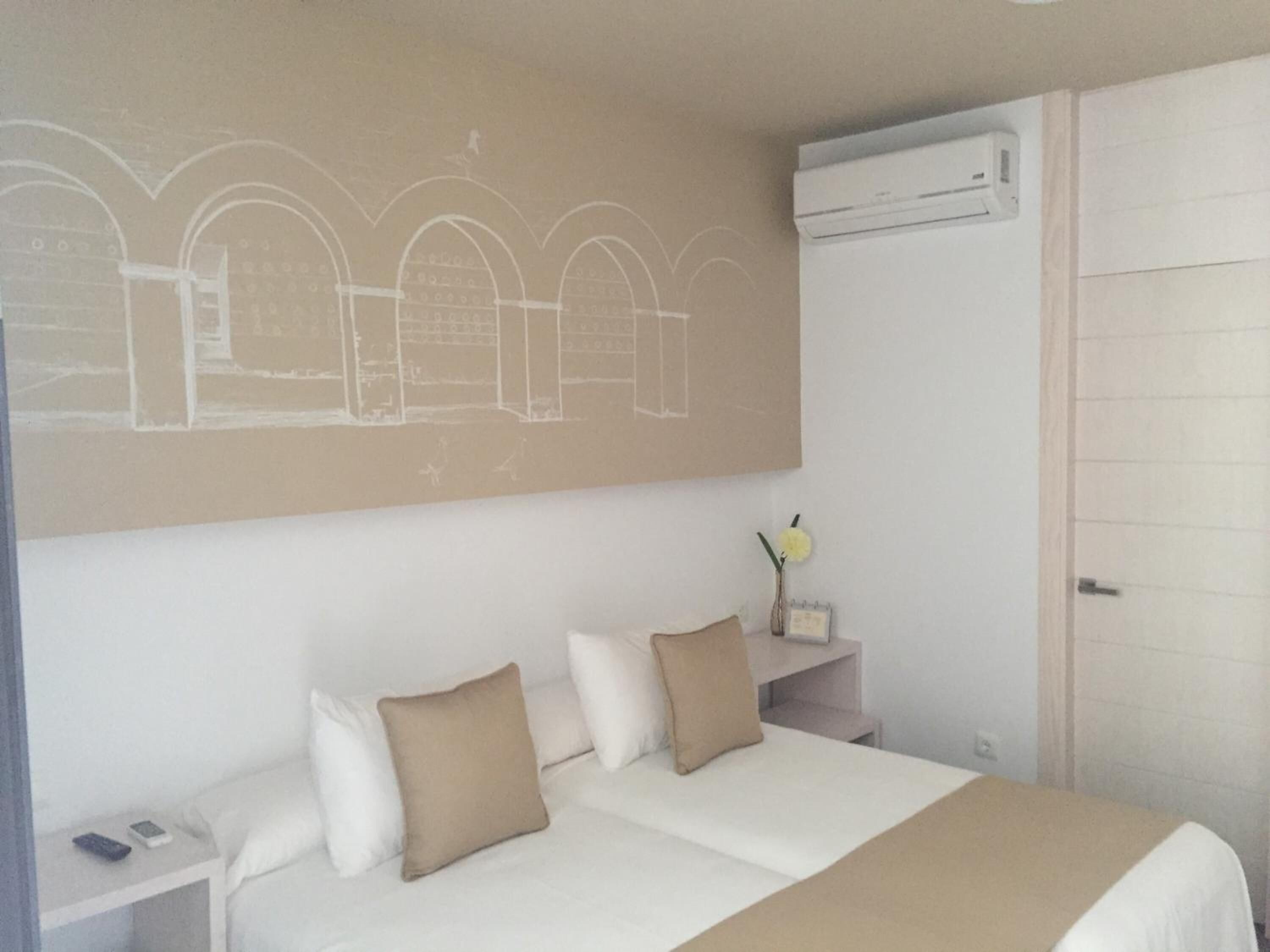 Una habitación de hotel con una cama, dos mesitas de noche, un aire acondicionado de pared y una puerta.