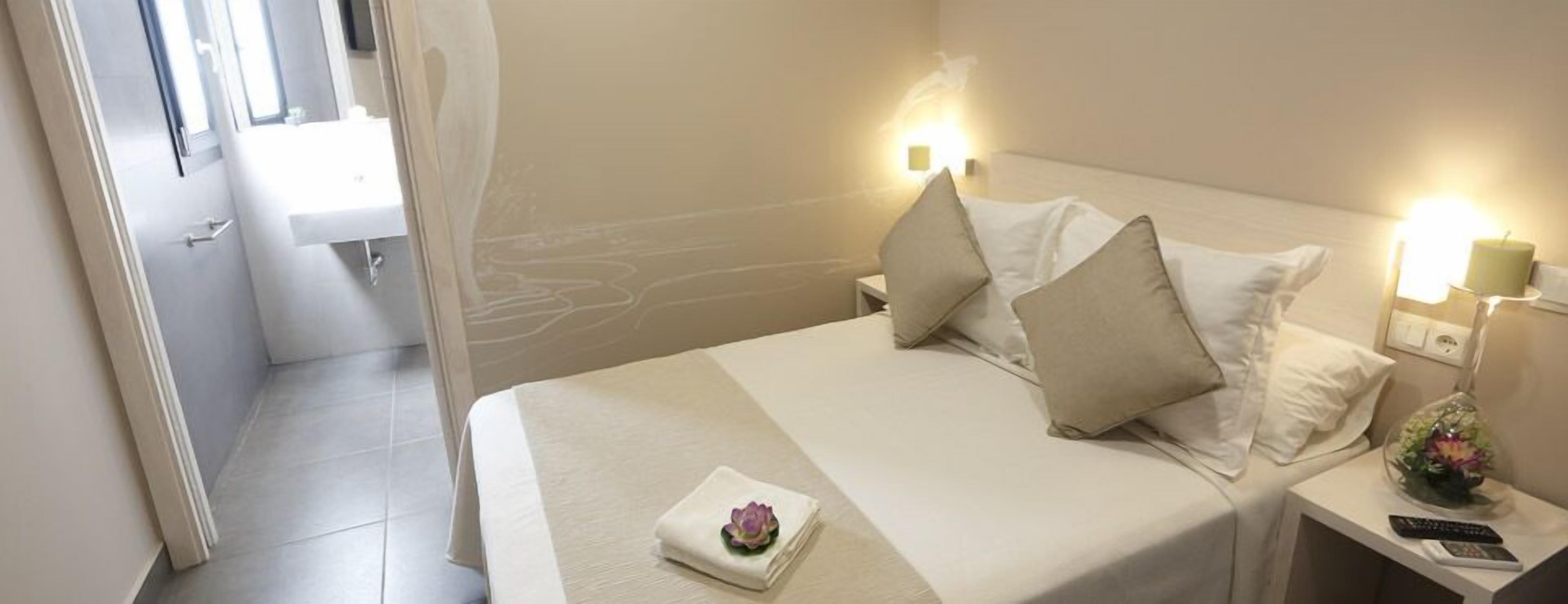 A hotel room with a bed, bedside tables, a nightstand with a lamp, a remote control, and a flower arrangement.
