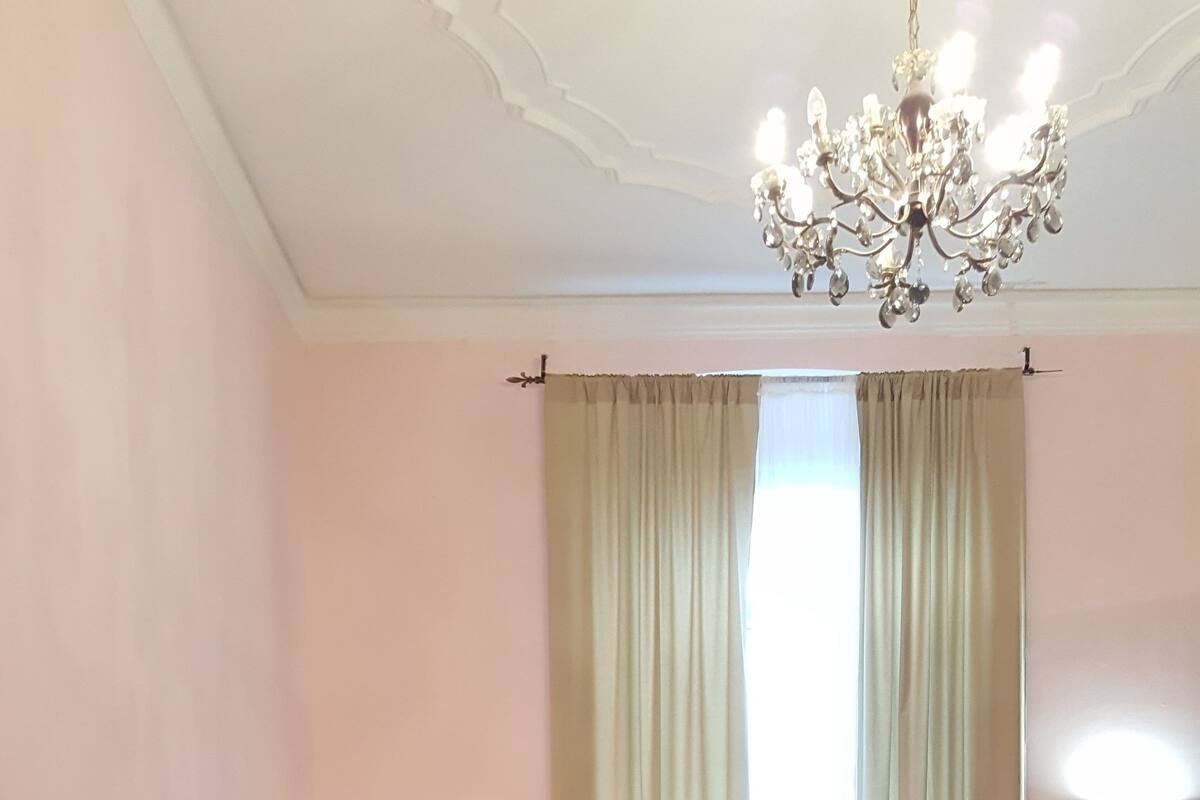A neatly arranged bedroom with a bed, pillows, a nightstand, a chandelier, and a window with curtains.