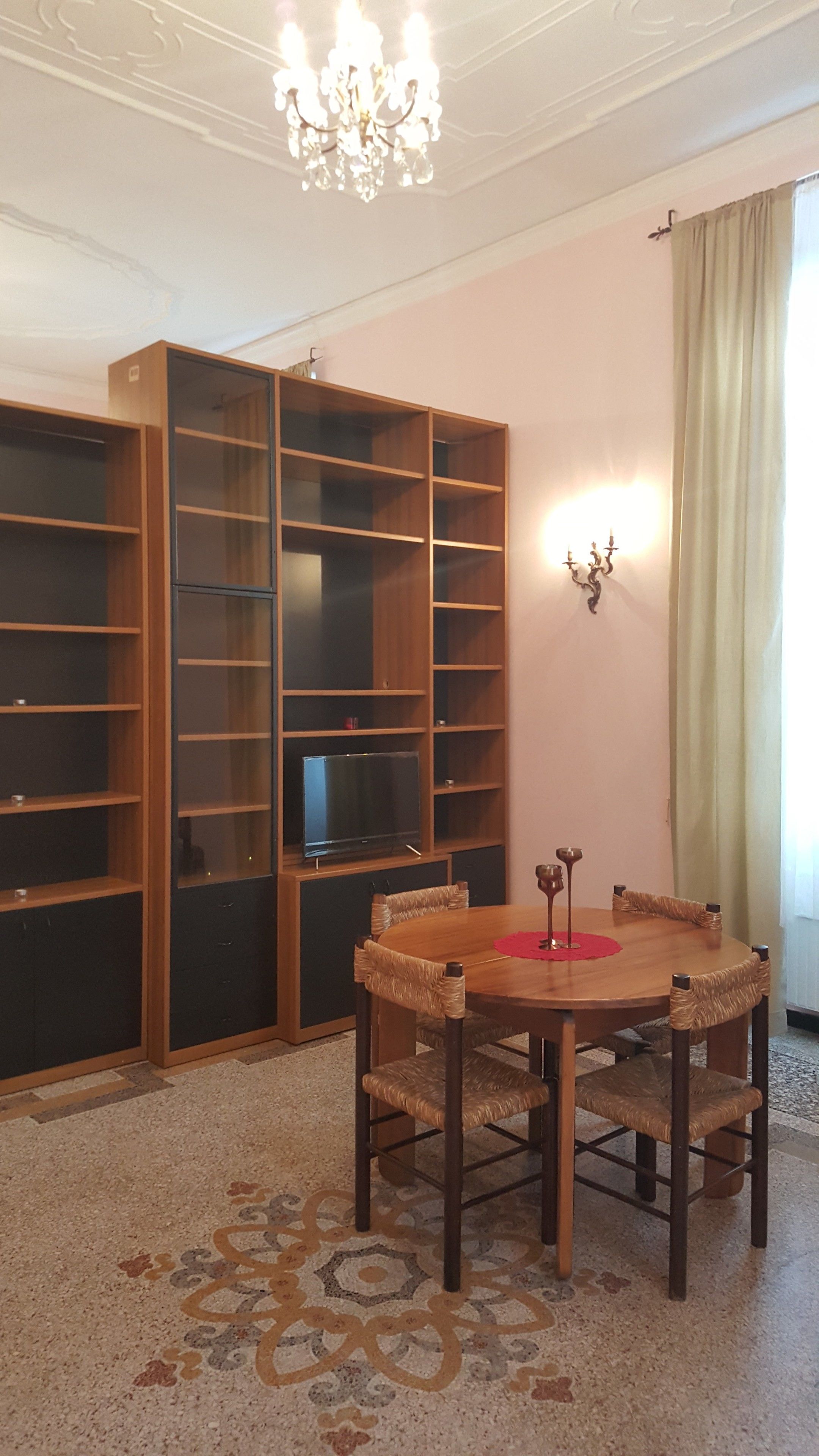 Une pièce comprenant une table à manger en bois, quatre chaises en osier, un lustre, un téléviseur posé sur un meuble et des étagères intégrées en bois.