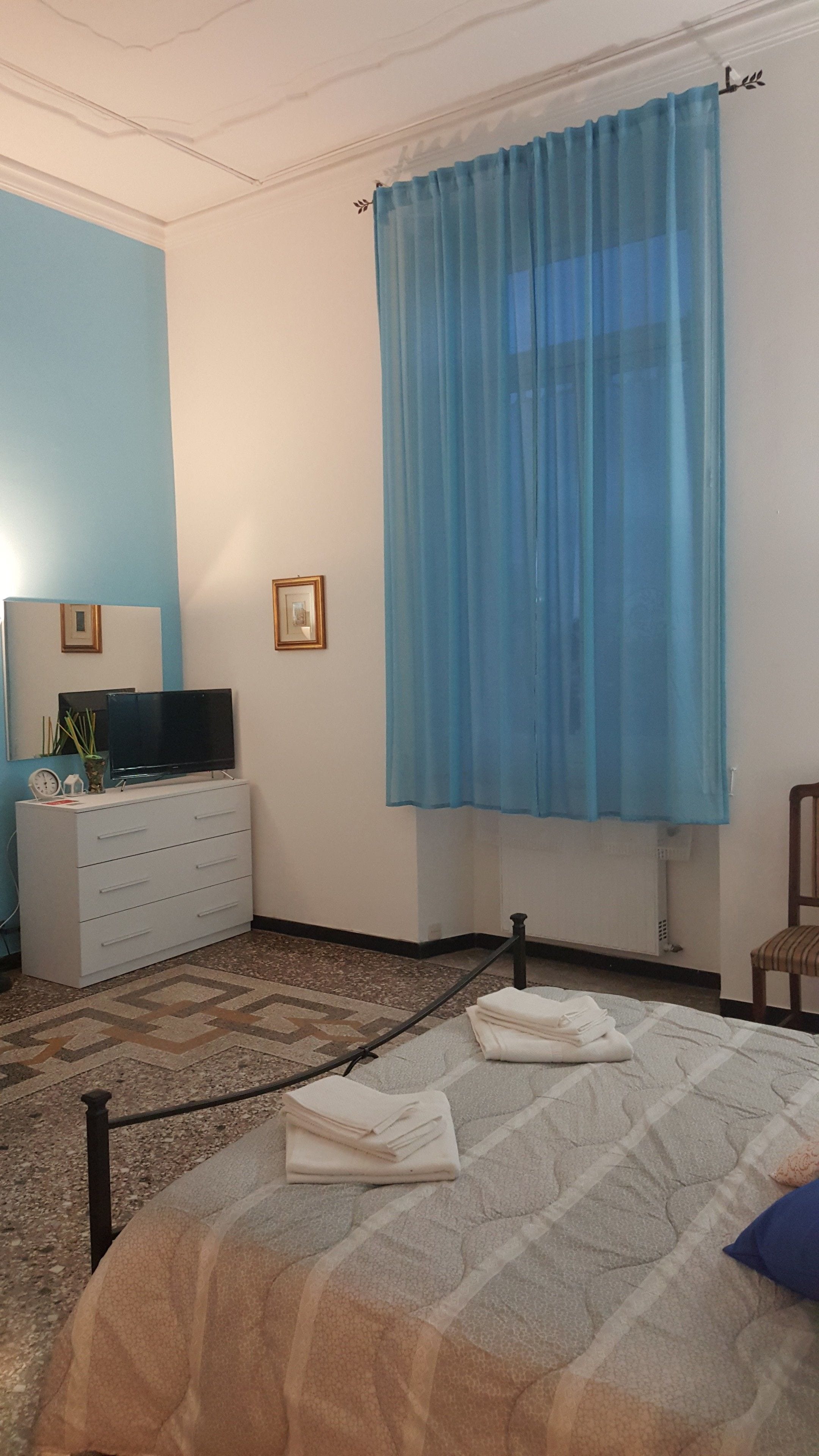 A bedroom with a bed, a TV on a dresser, a window with blue curtains, and a small chair.