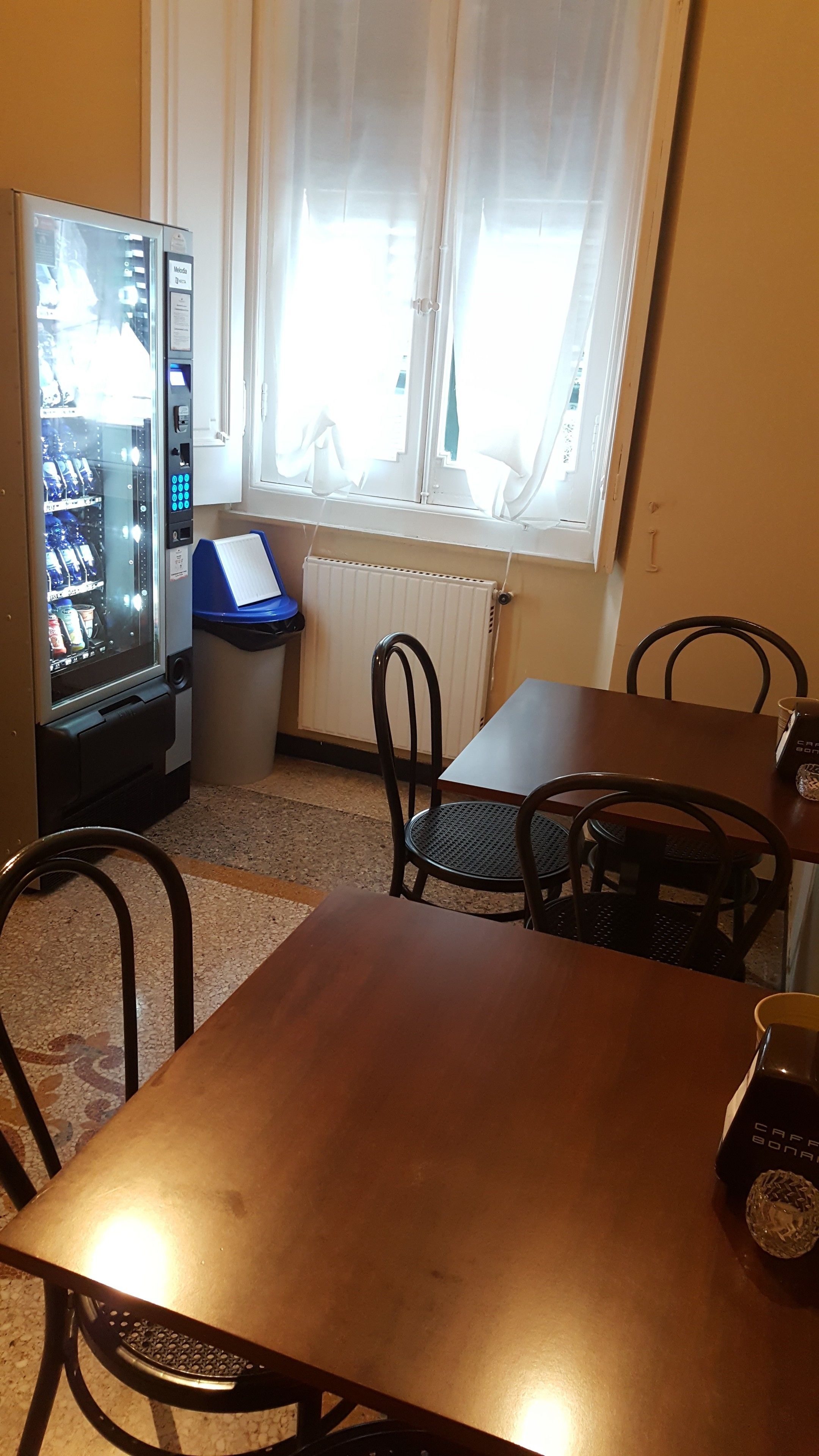 A room with a vending machine, a window with curtains, a radiator, a table, and chairs.