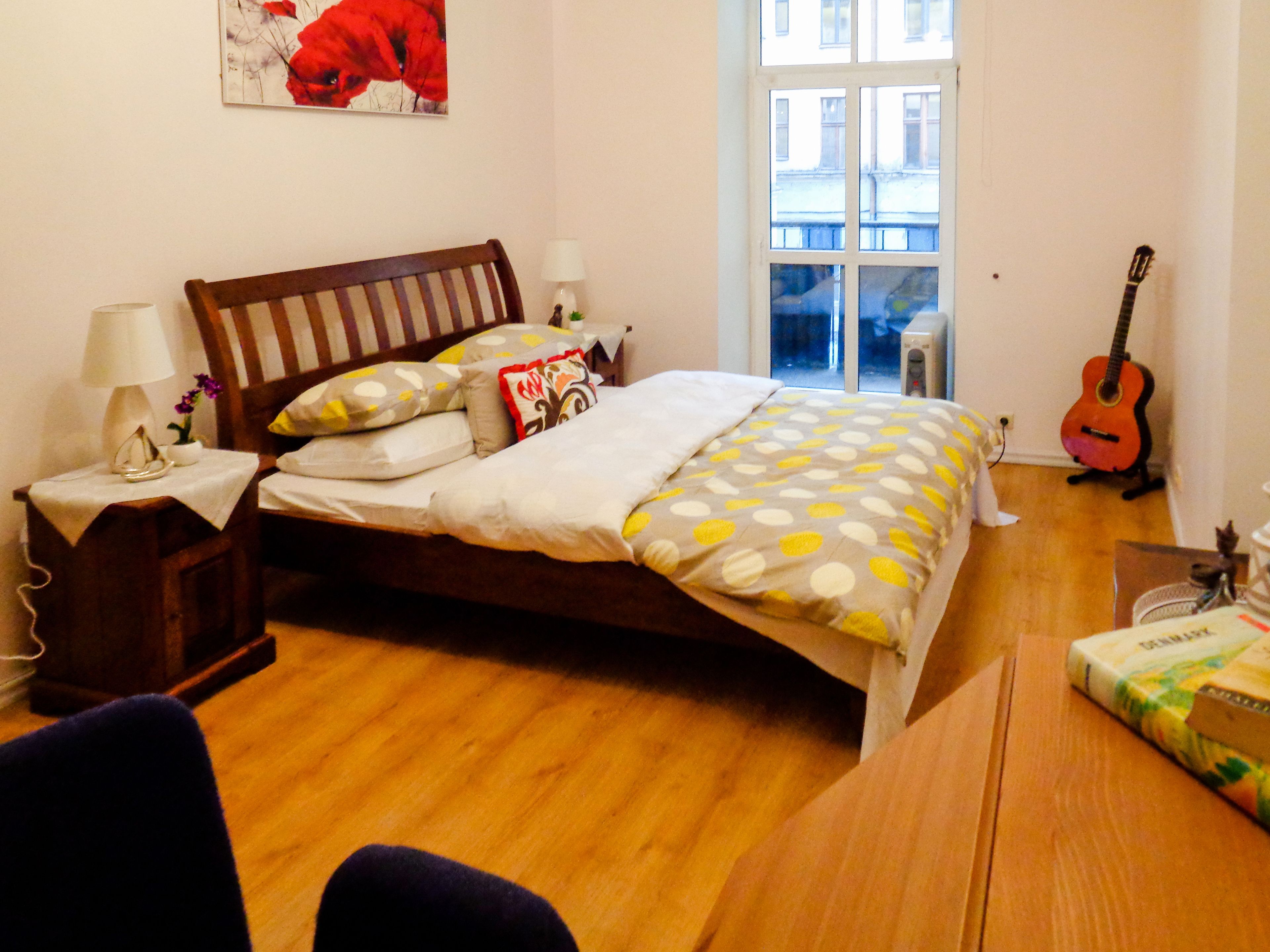 A bedroom with a wooden bed, a nightstand, a lamp, a guitar, and a window with a view of buildings.