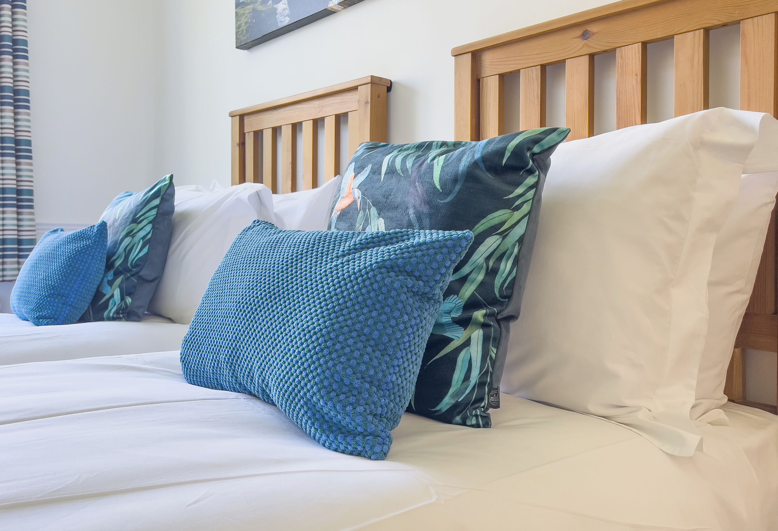 Cama con ropa de cama blanca, almohadas azules, cabecero de madera y un cuadro colgado en la pared.