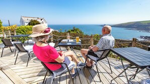 Two people seated at a wooden deck with a view of the sea and a white building in the background. - Mevagissey Bay Hotel (St Austell)