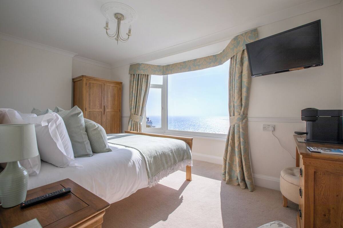 A bedroom with a bed, a TV, a desk, and a view of the sea.