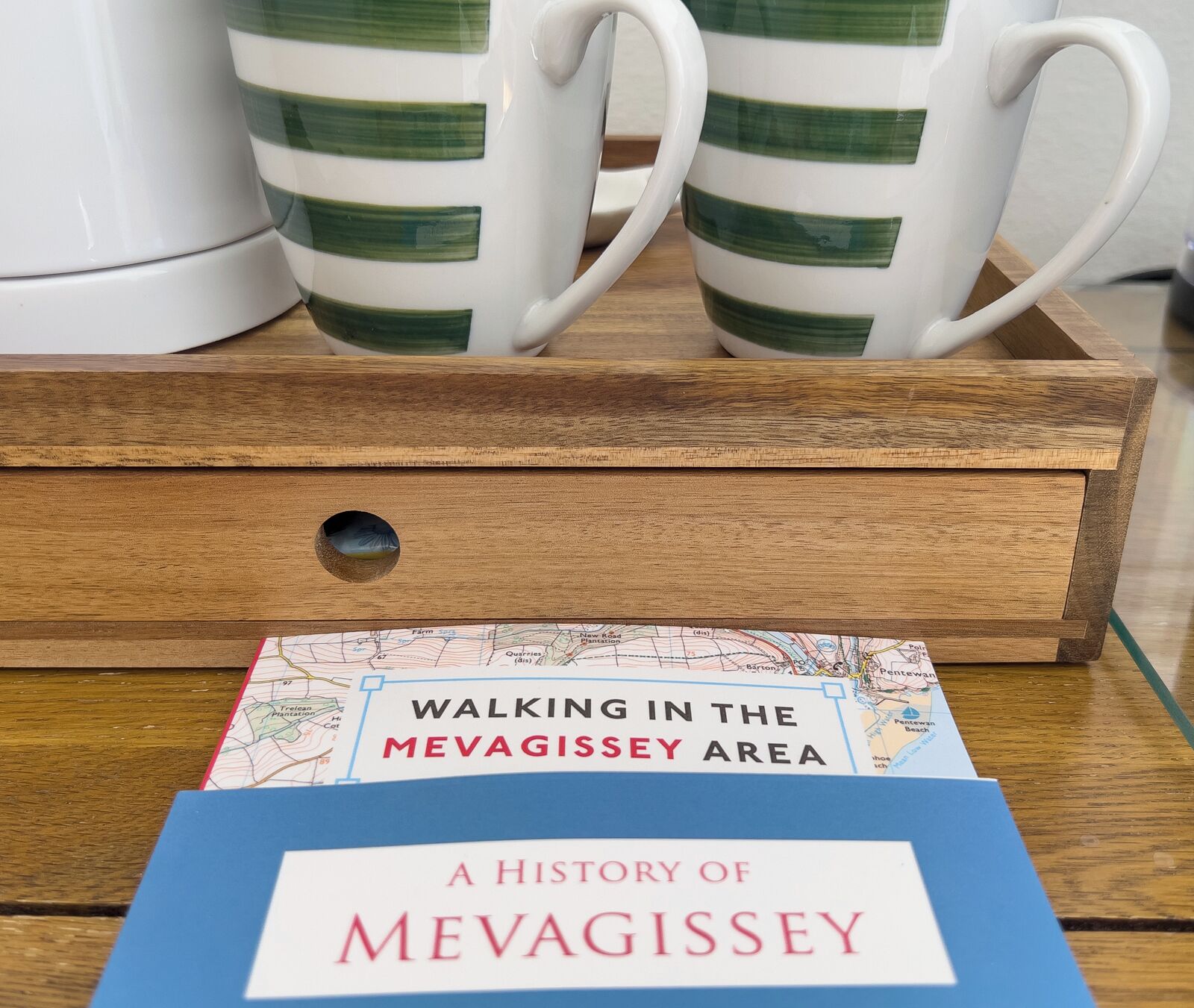 A wooden tray with two striped mugs and a book titled "A History of Mevagissey" on a wooden table.