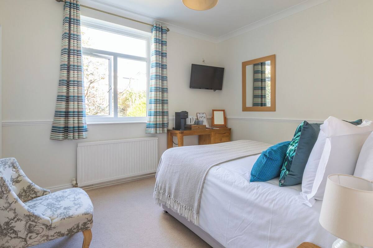 A bedroom with a bed, a chair, a bedside table, a TV, and a window with curtains.