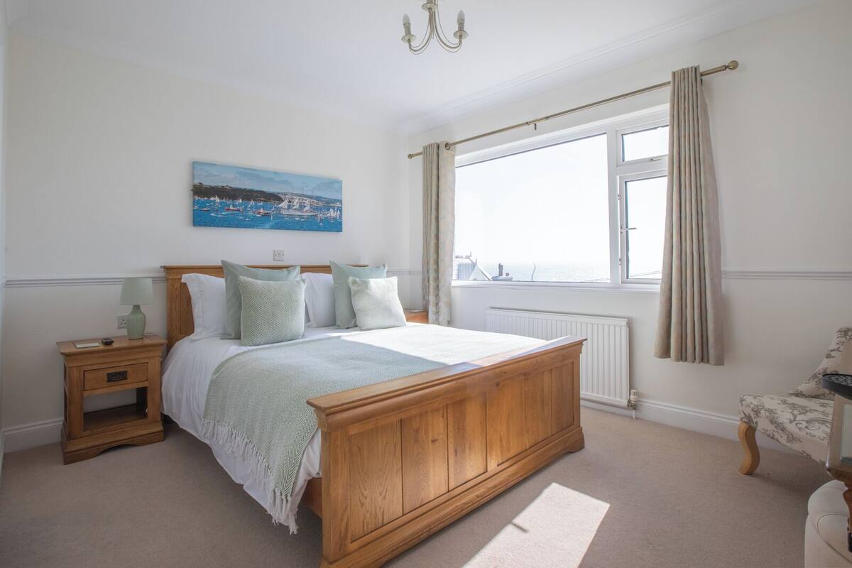 A bedroom with a wooden bed, a nightstand, a bedside table, a window with curtains, and a painting on the wall.