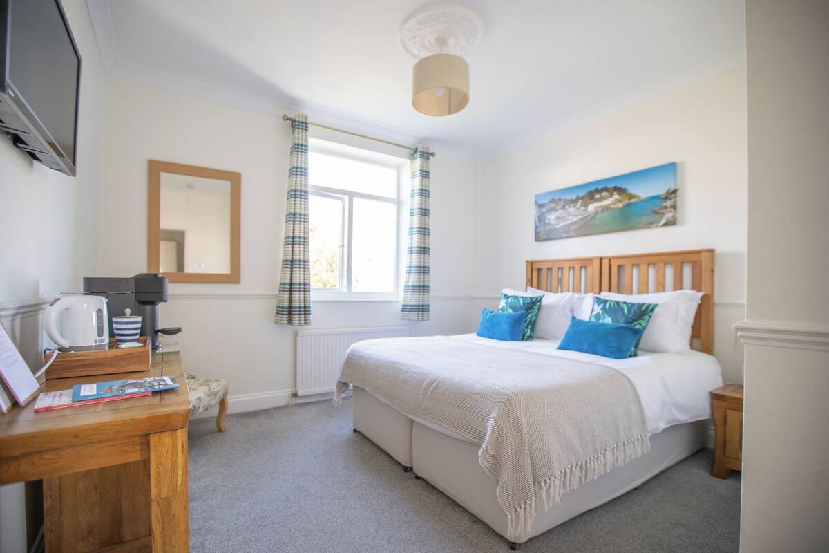 A bedroom with a bed, a bedside table with a kettle, a coffee maker, and a framed picture on the wall.