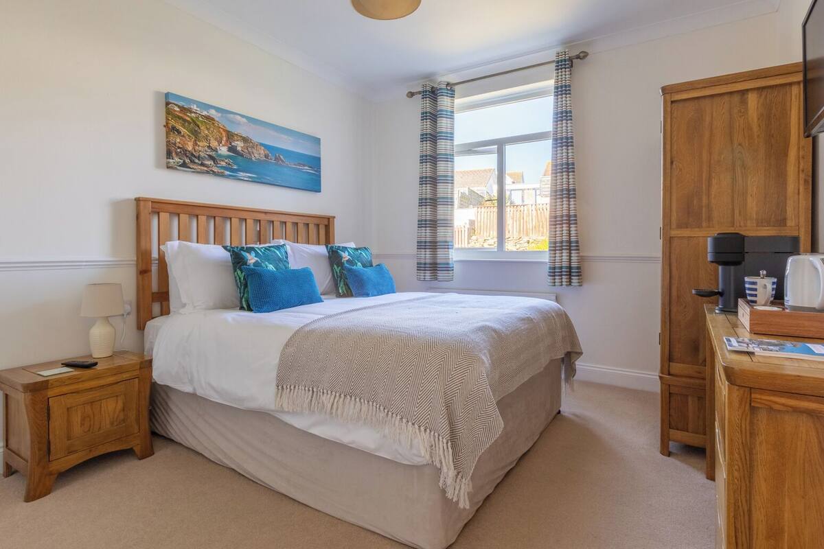 A bedroom with a wooden bed, a nightstand, a bedside table, a window with curtains, a painting on the wall, and a wardrobe.
