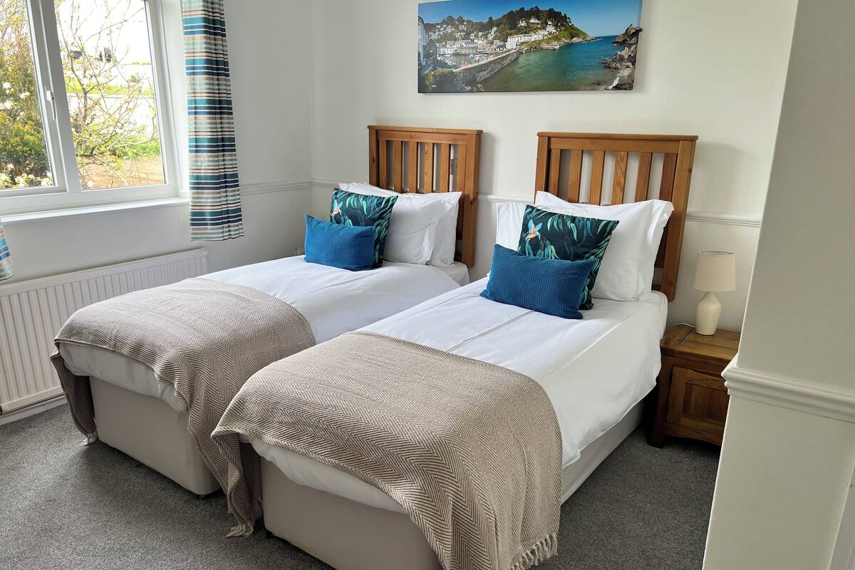 A double room with two beds, each with a wooden headboard, white bedding, and blue patterned pillows. There is a window with curtains, a radiator, and a painting of a coastal scene on the wall.