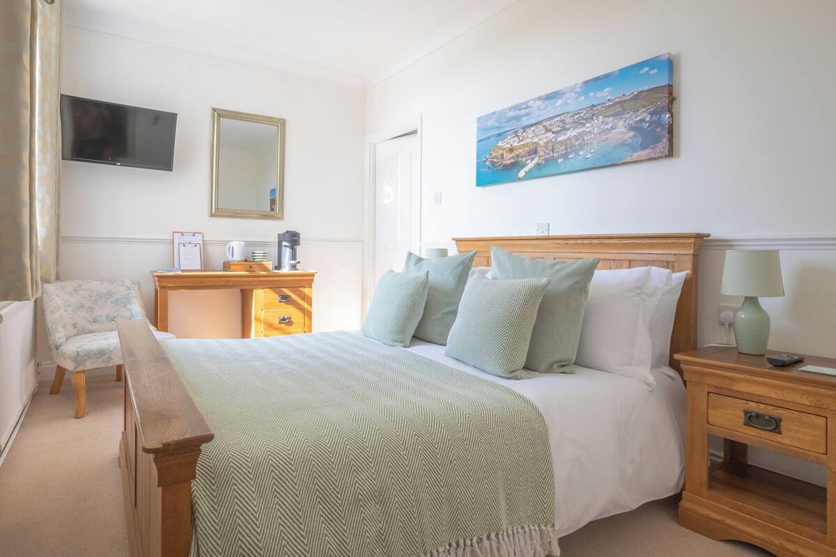 A hotel room with a bed, a desk, a chair, a TV, a painting, and a nightstand with a lamp.