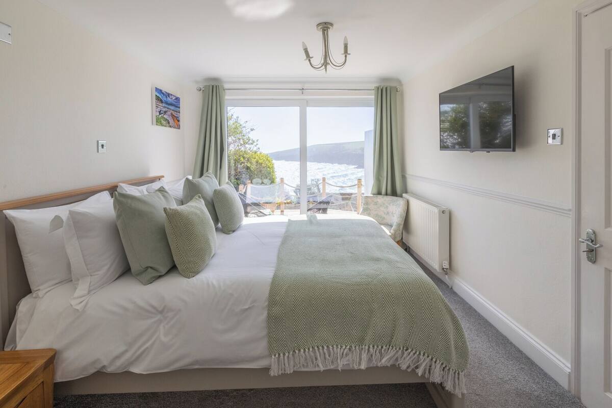A bedroom with a bed, a television, a window with curtains, and a view of the outdoors.