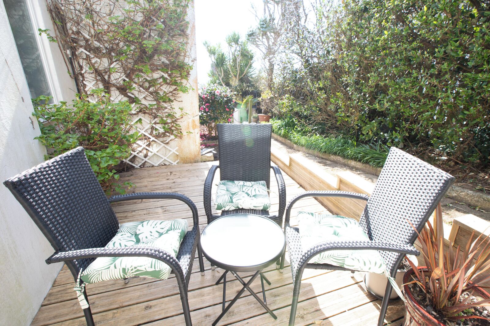 A patio with three wicker chairs and a table, surrounded by potted plants and a wooden deck.