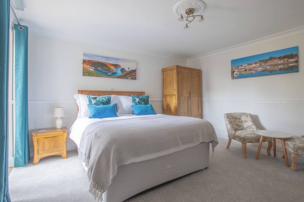 A bedroom with a large bed, a wooden wardrobe, a chair, and two paintings on the walls.
