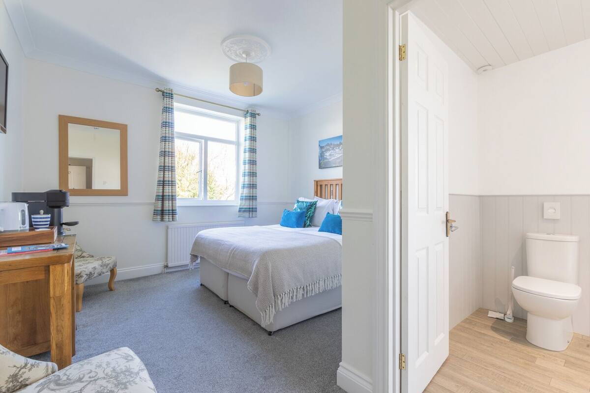 A bedroom with a bed, a bedside table, a chair, a window with curtains, a door, and a toilet.