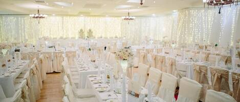 Un salón de banquetes con filas de mesas dispuestas para un evento formal, adornadas con manteles blancos y sillas, y decorado con centros de mesa florales.