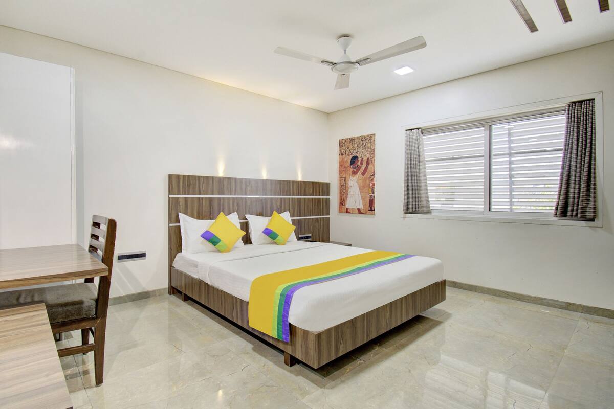 Quarto de hotel com cama, escrivaninha, cadeira, ventoinha no teto, janela com persianas e um quadro na parede.