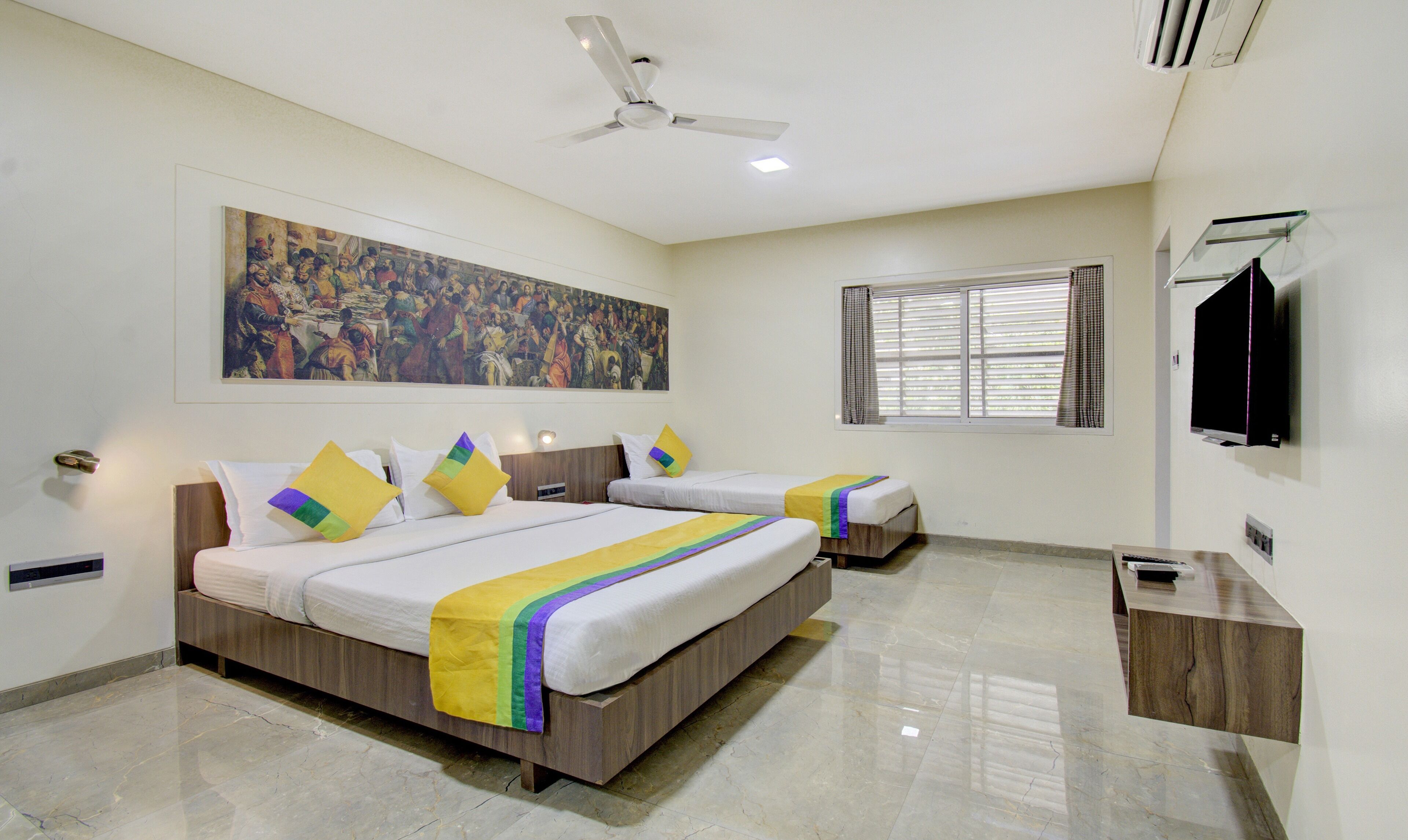 A hotel room with two beds, a ceiling fan, a large painting on the wall, a window with curtains, and a flat-screen TV mounted on the wall.