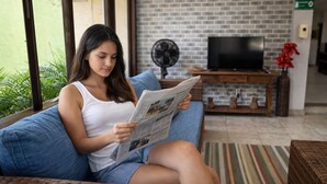 Una persona sentada en un sofá leyendo un periódico en una habitación con pared de ladrillo, un televisor y un ventilador.