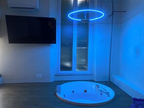 Ett rum med ett stort fönster, en väggmonterad TV och ett runt badkar med en blå lampa ovanför.