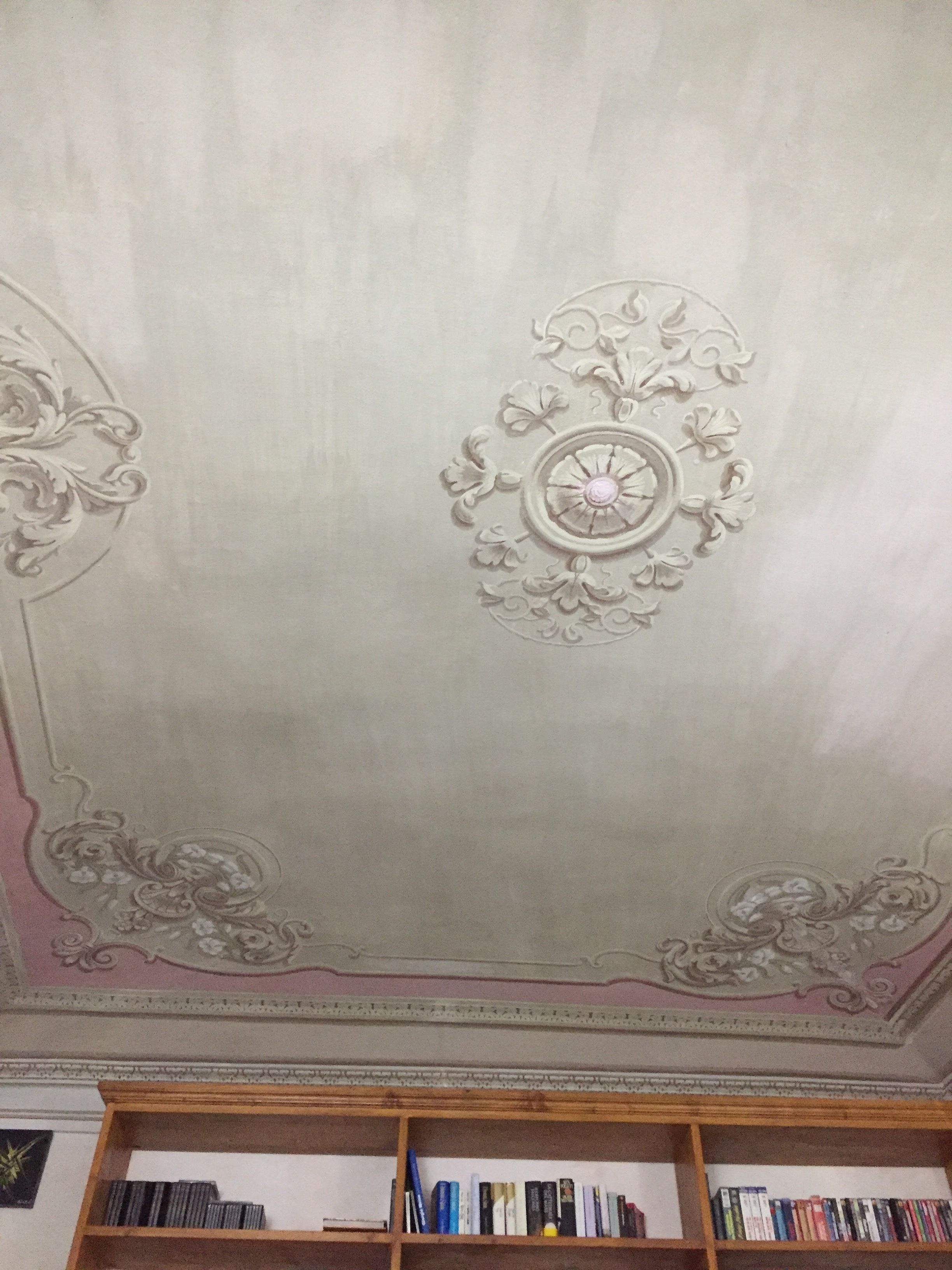 A ceiling with ornate floral and geometric patterns, a wooden bookshelf below, and a plain wall.