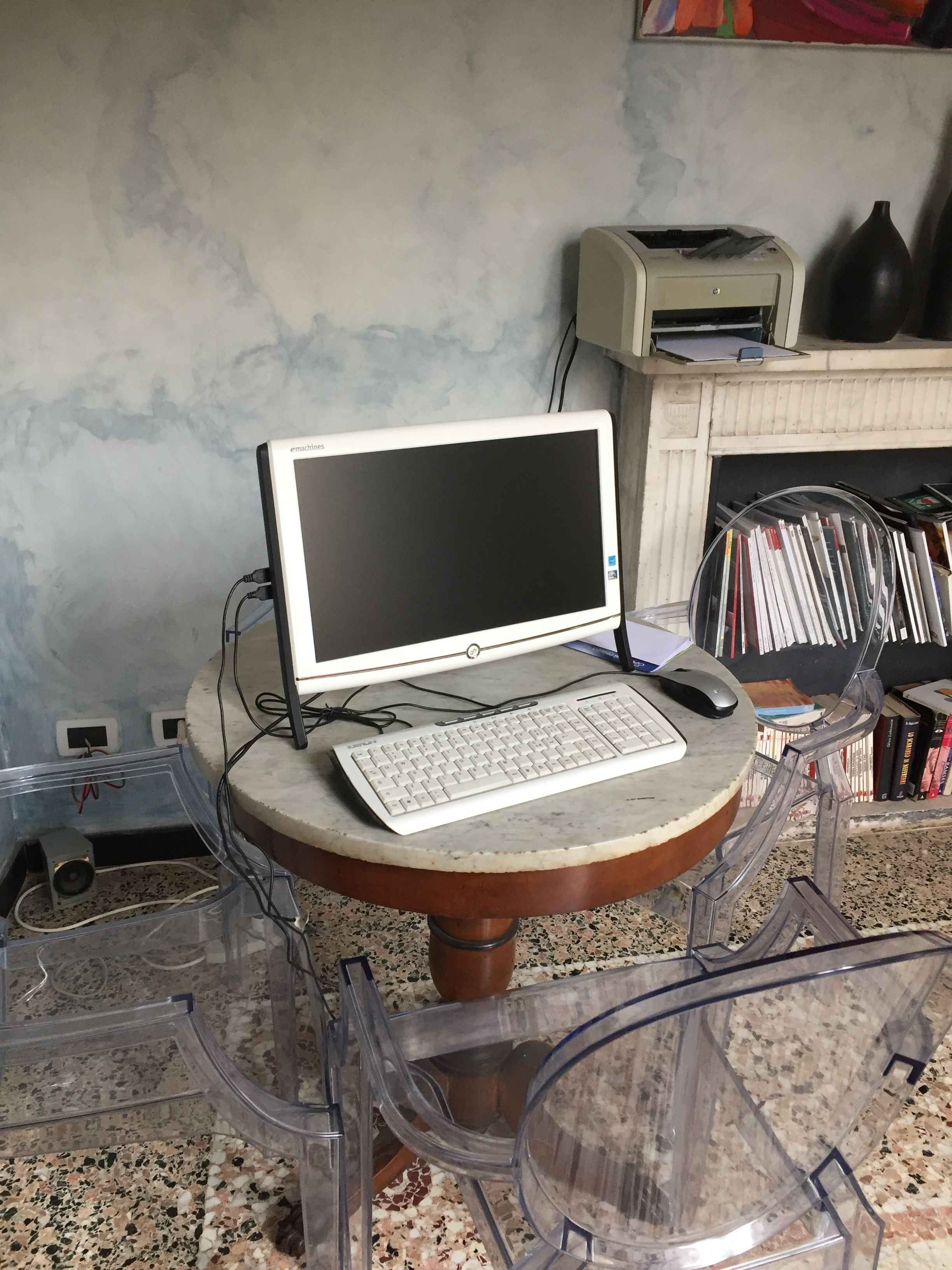 A desktop computer on a round table with a keyboard and mouse, surrounded by clear plastic chairs.
