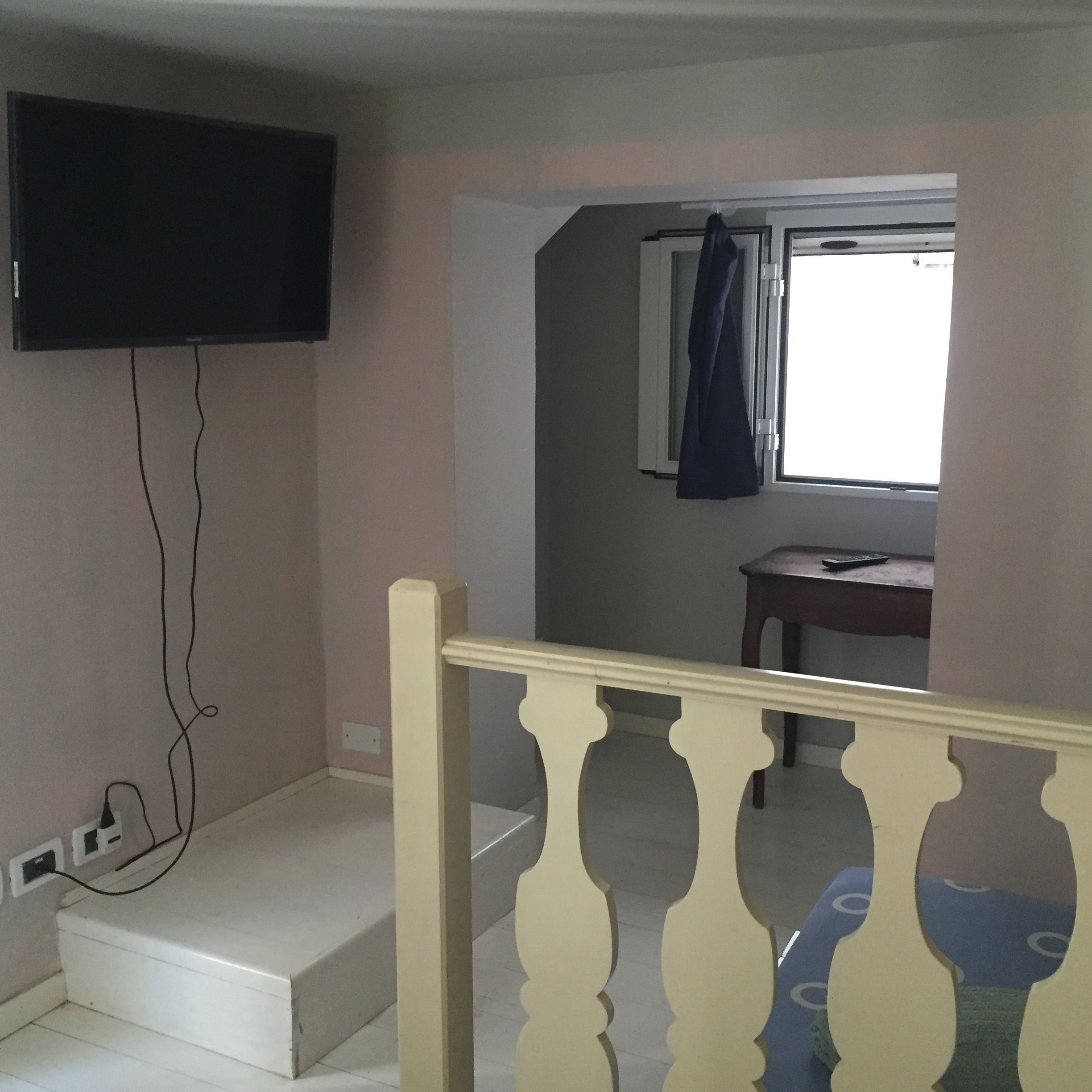 Ein Zimmer mit einem an der Wand befestigten Fernseher, einem Fenster mit Vorhang, einem kleinen Tisch und einer Treppe mit Geländer.