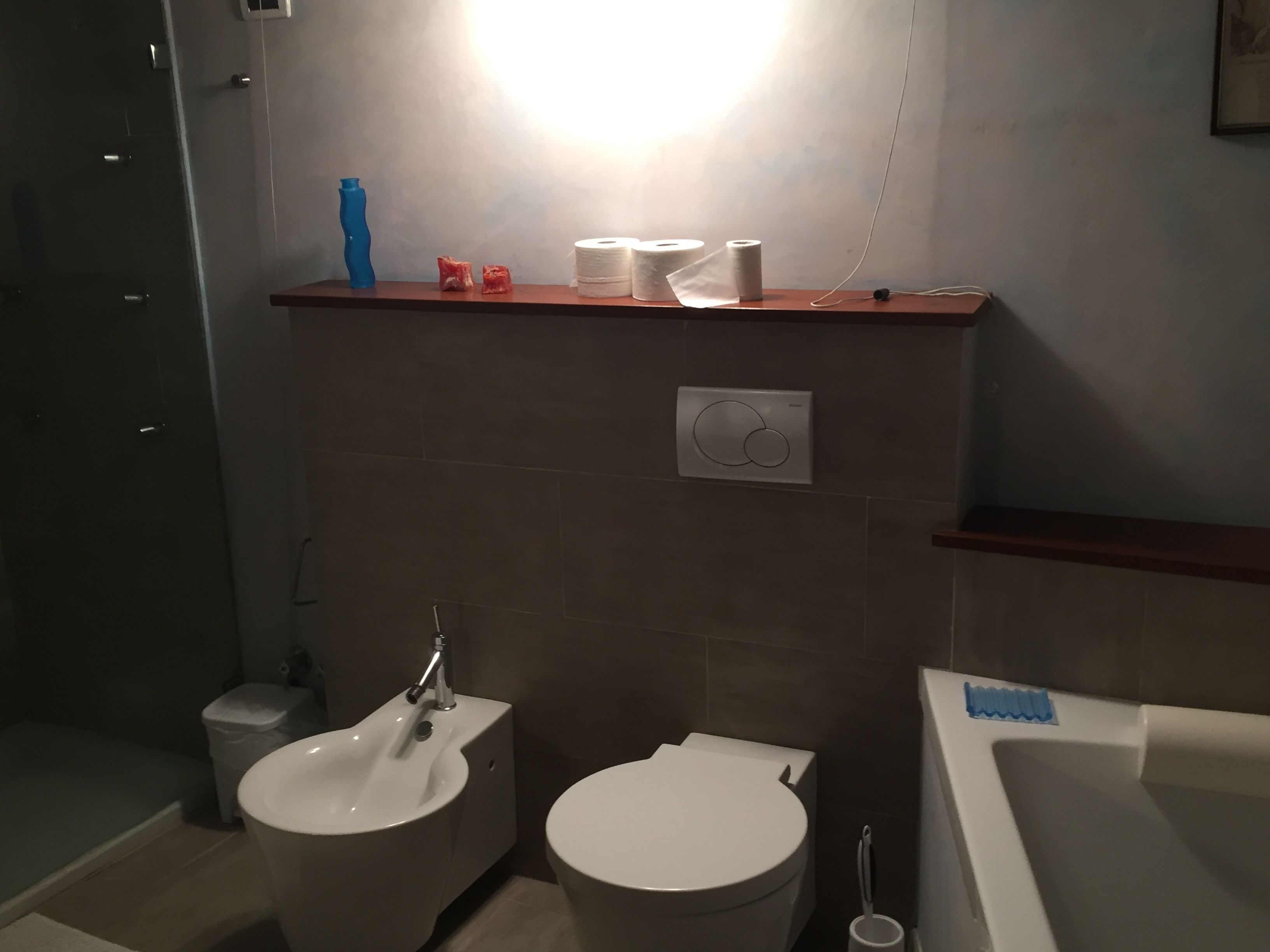 Un baño con lavamanos, inodoro y bañera. Hay un estante con papel higiénico y una botella azul.