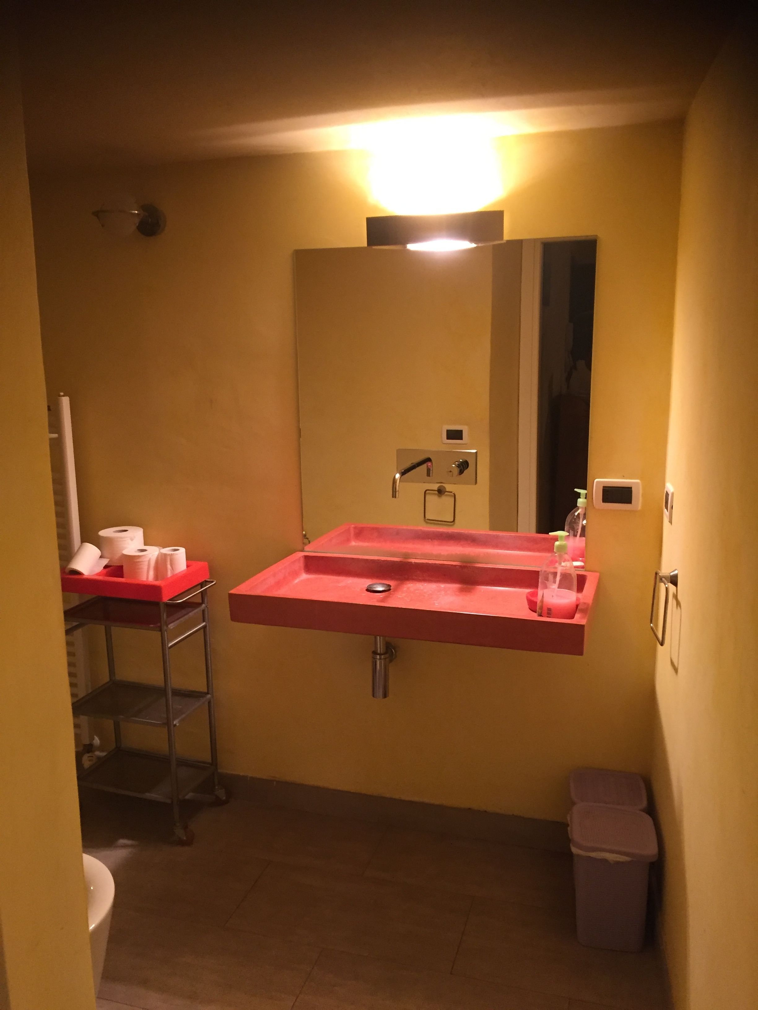 Une salle de bain avec un lavabo rouge, un miroir et des toilettes. On y trouve une poubelle et une étagère avec des articles de toilette.