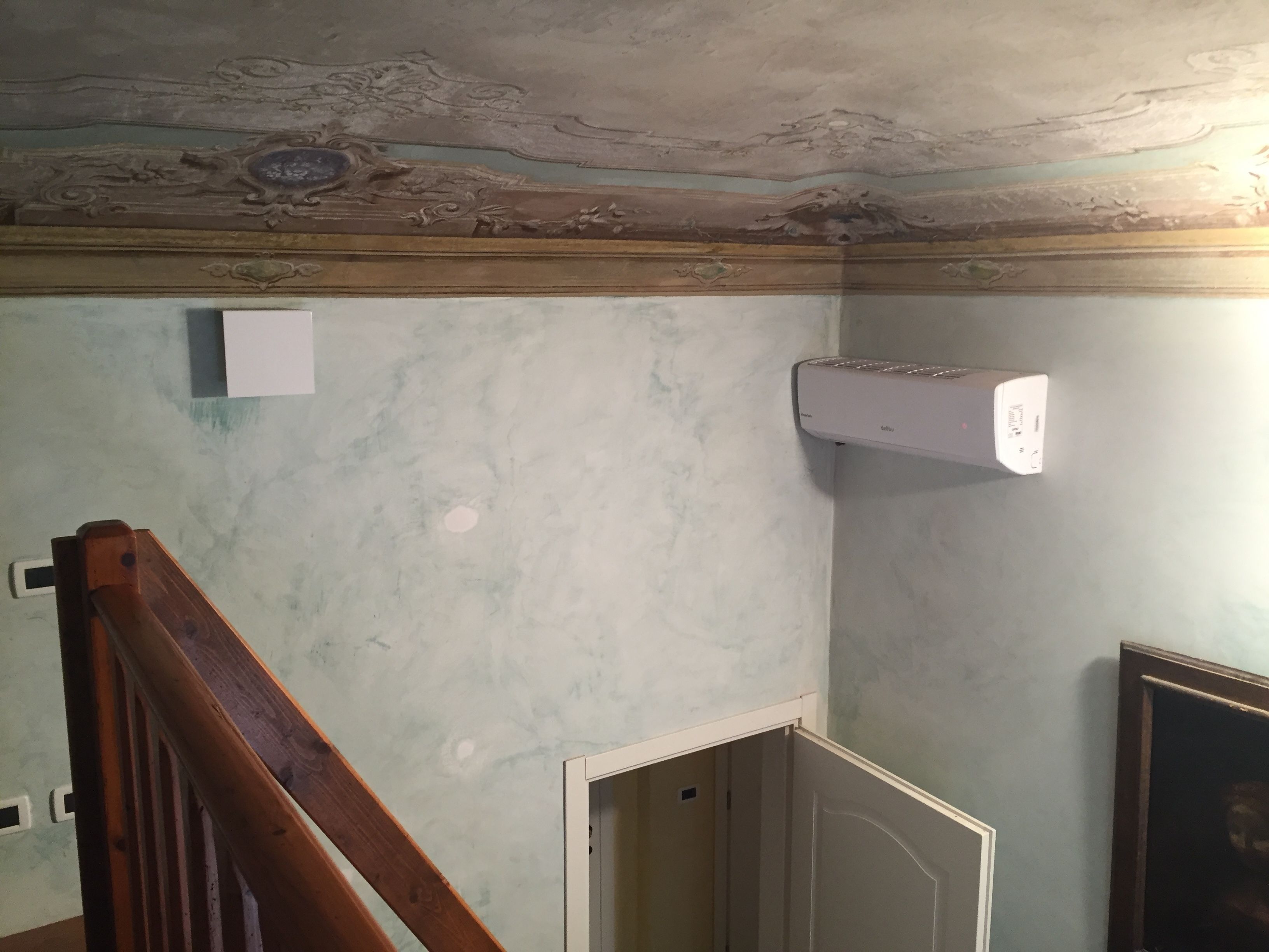 A room with a wooden staircase, a white air conditioning unit mounted on the wall, and a partially painted wall with decorative ceiling details.