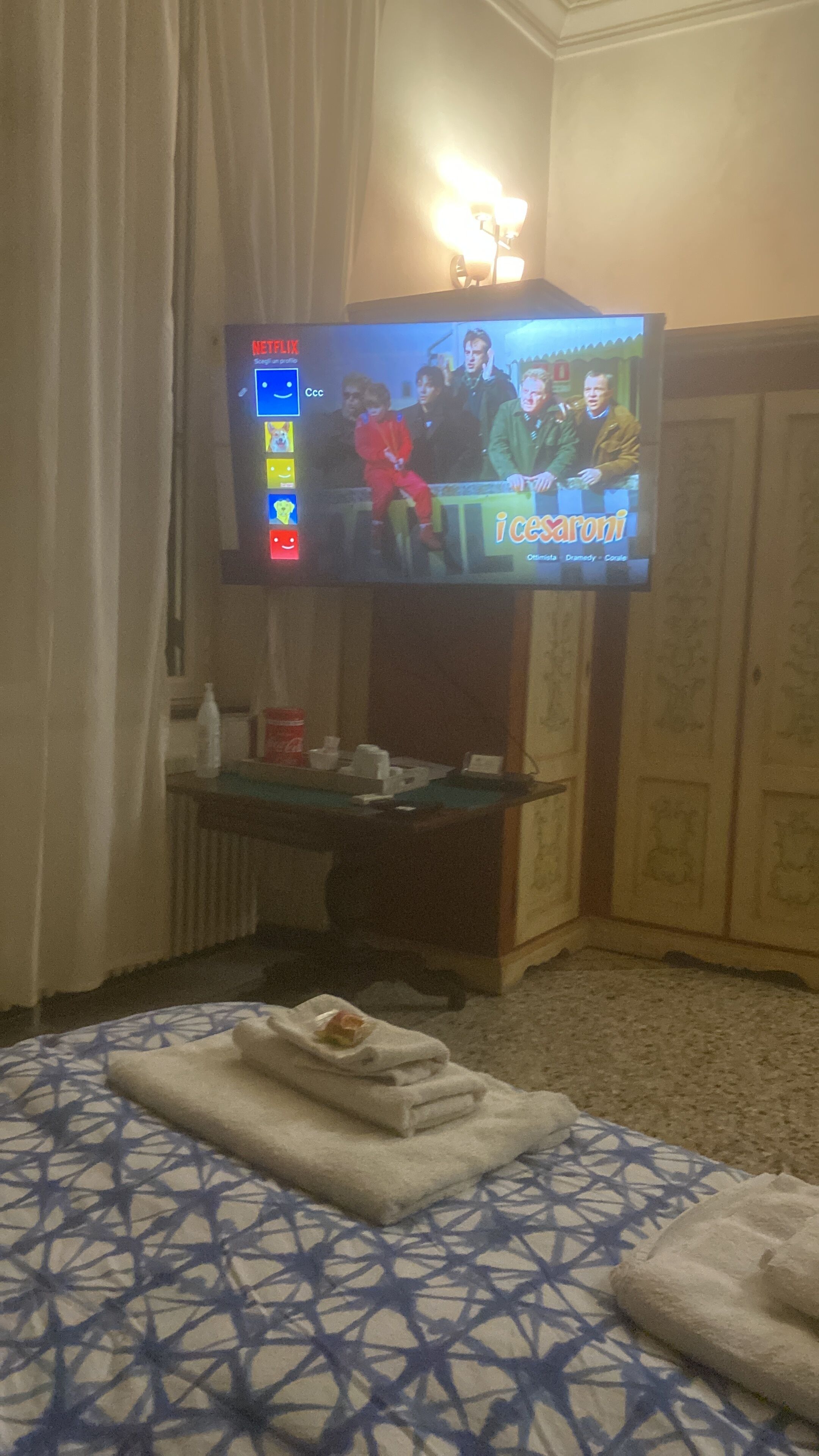 A hotel room with a television on a stand, a small table with a bottle and glasses, and a bed with a patterned bedspread.
