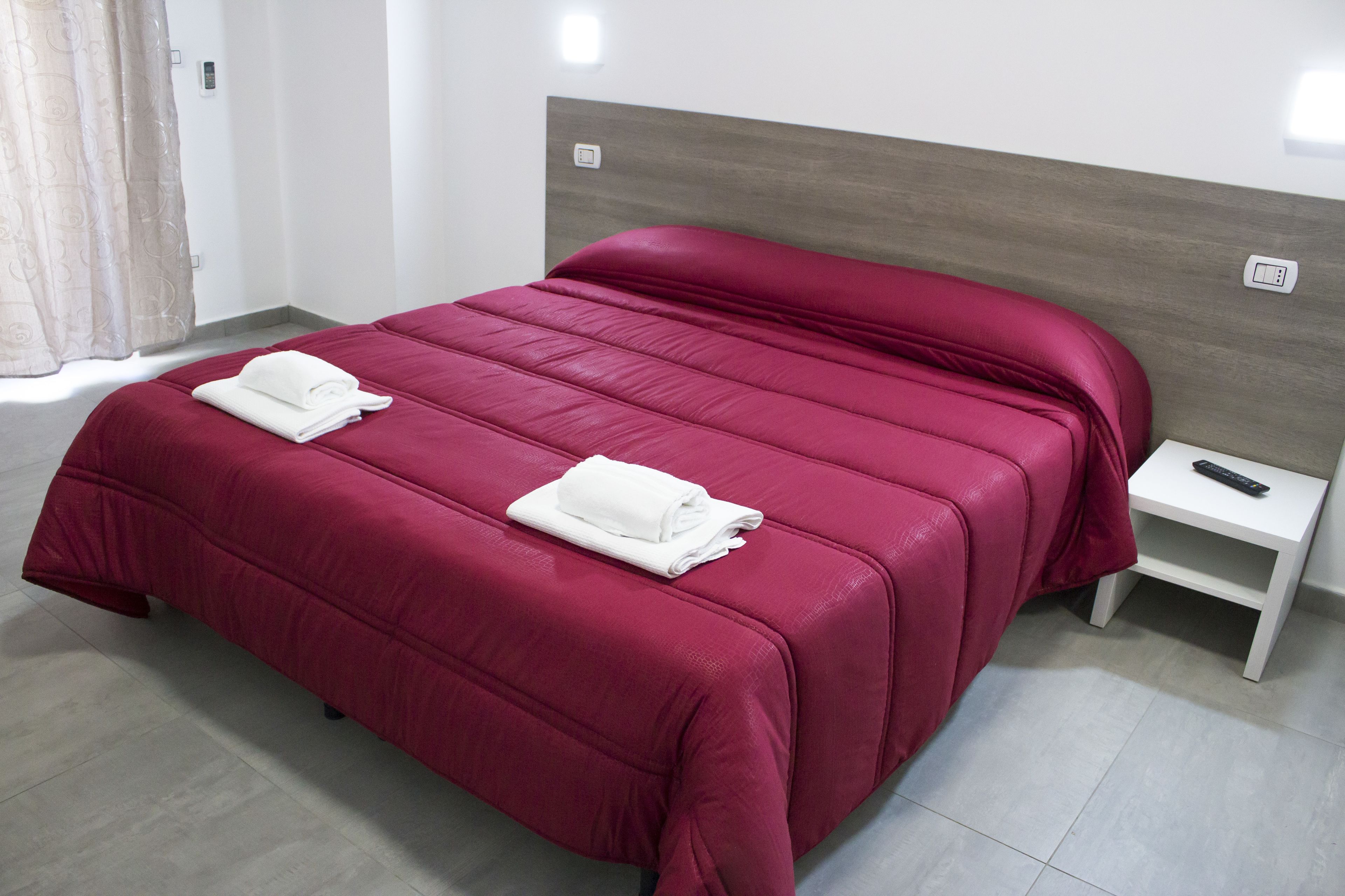 Una cama bien tendida con una colcha roja y dos toallas blancas dobladas, una mesita de noche blanca con un control remoto y un interruptor de luz en la pared.