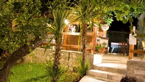 Un espacio exterior bien iluminado con escalones que conducen a una estructura de madera, rodeado de vegetación y un muro de piedra.