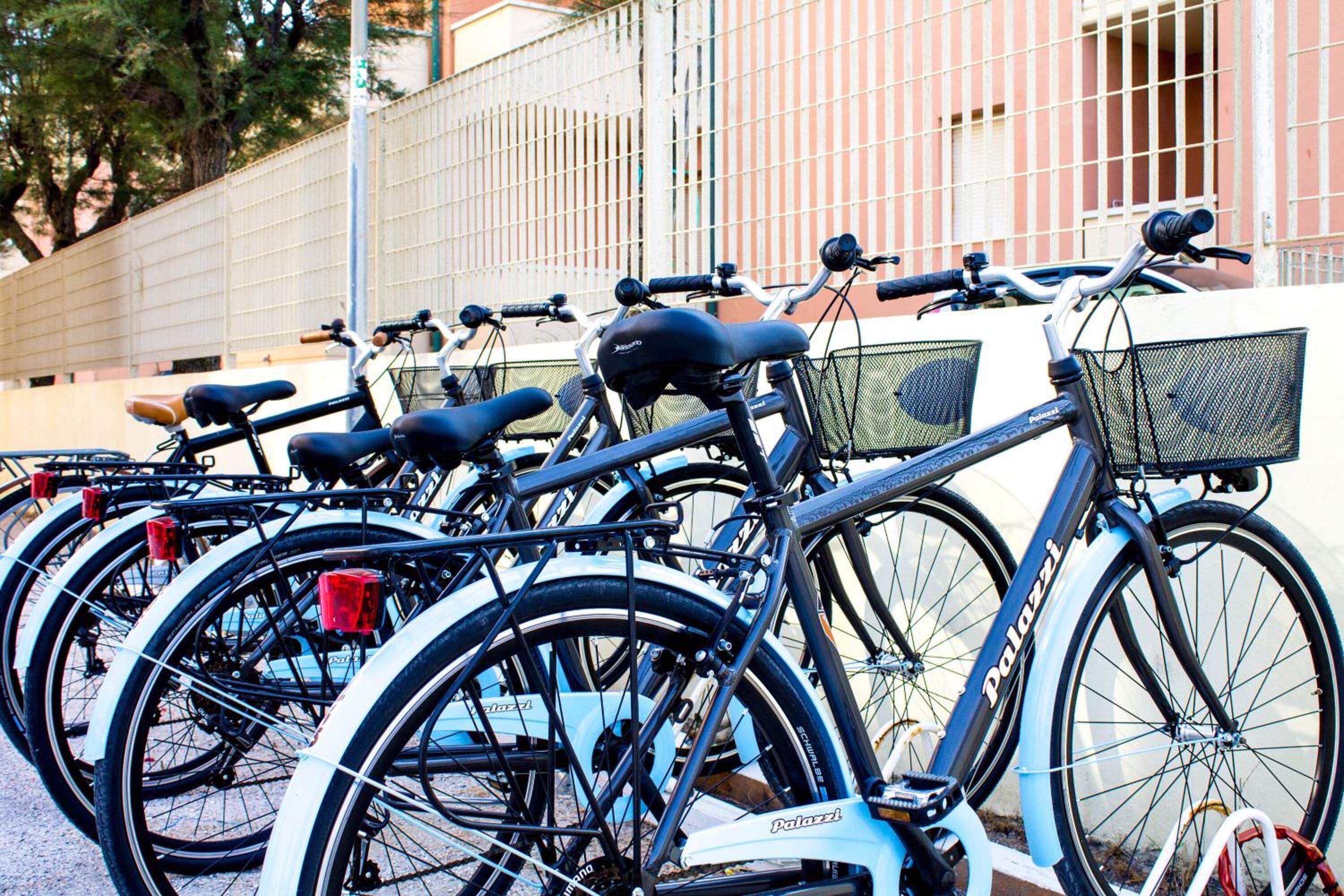 Várias bicicletas estacionadas em frente a um prédio com cerca de metal e árvores ao fundo.