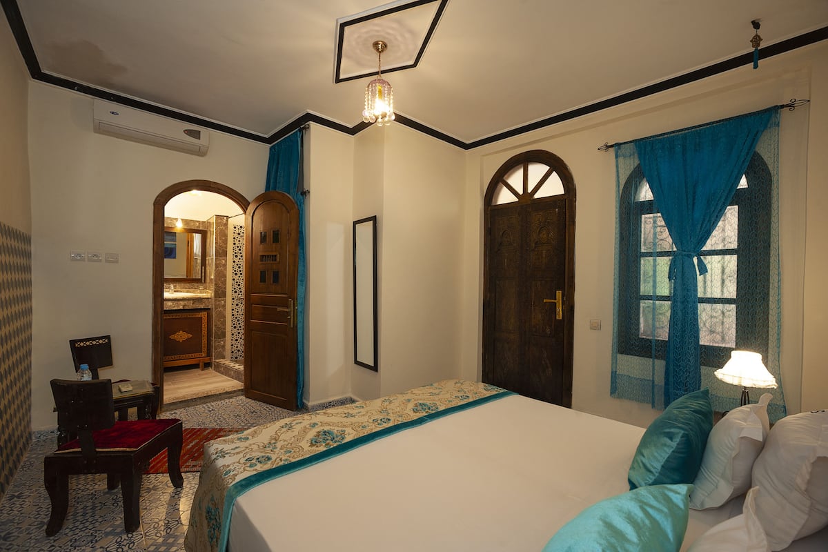 Une chambre avec un grand lit, une armoire en bois, un bureau avec une chaise, et une salle de bain visible à travers une arche.