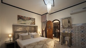 Un dormitorio con una cama grande, cabecera de madera, un cuadro sobre la cama, un espejo y un baño visible a través de un arco.
