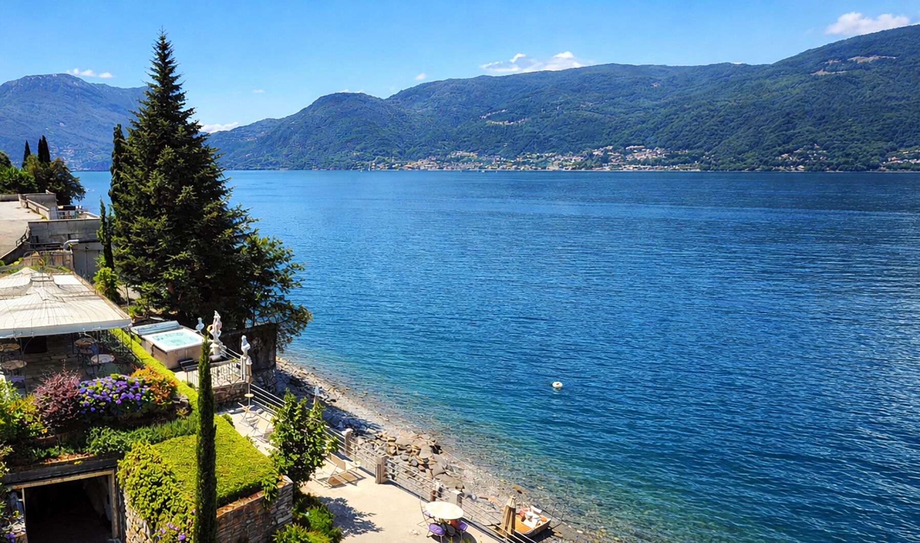 A lakeside view with a clear sky, mountains in the background, and a building with outdoor seating and a garden.